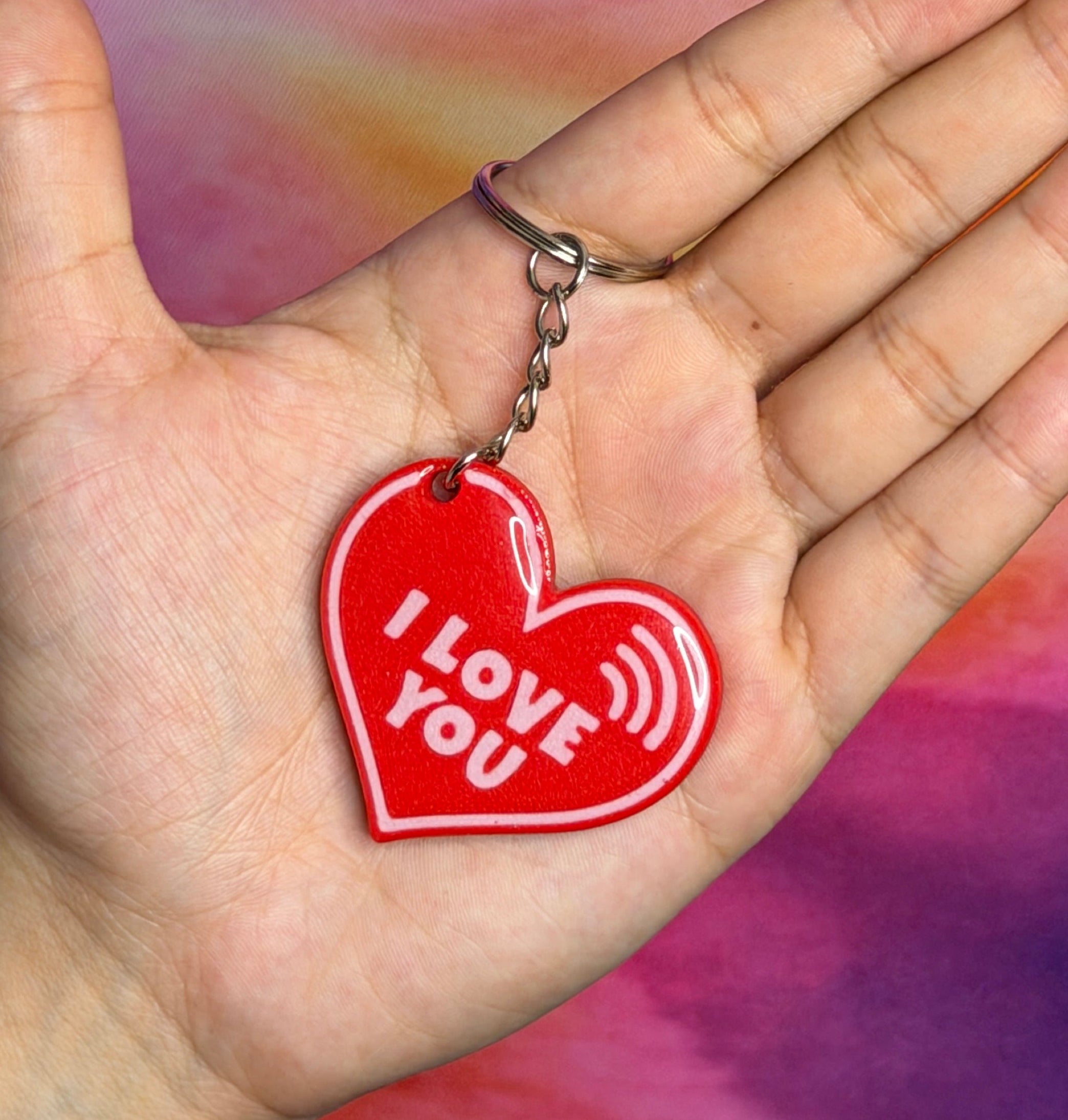 Hand holding a red heart-shaped keychain with 'I Love You' text against a colorful, abstract background.