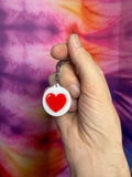Hand holding a keychain with a red heart design against a tie-dye background