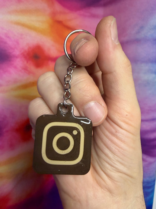 Hand holding a keychain with an  chocolate and creme Instagram logo against a colorful background